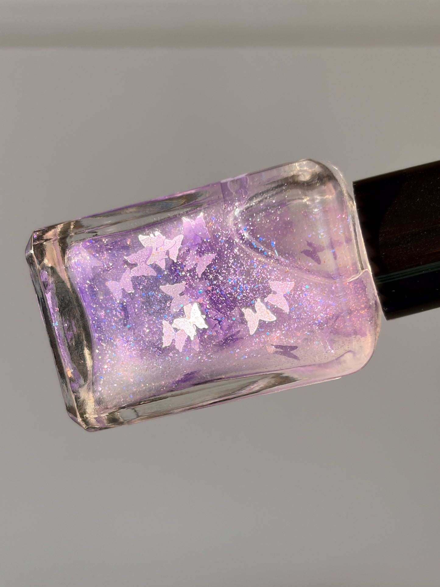 Nail polish bottle with purple glitter and butterfly design on a gray background