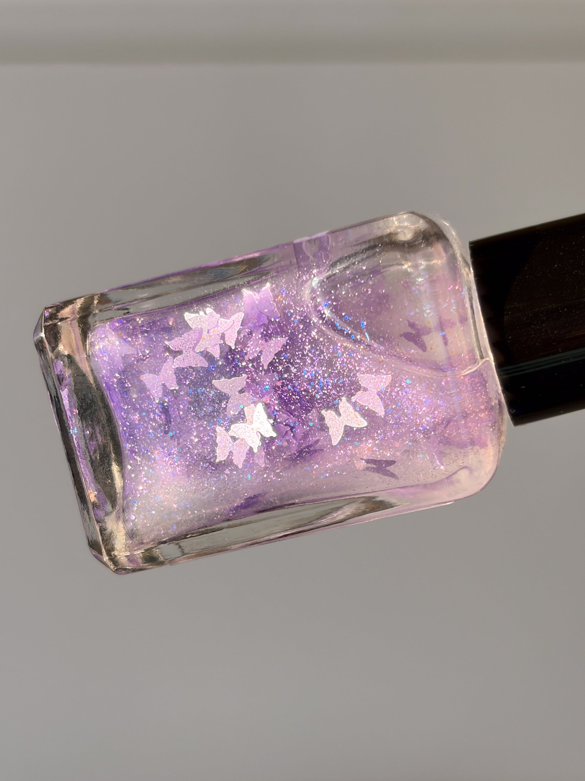 Nail polish bottle with purple glitter and butterfly design on a gray background