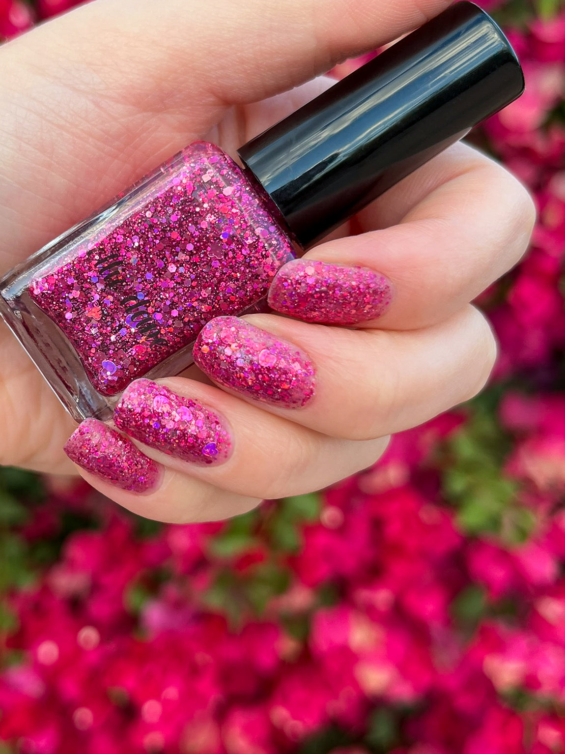 Hand with pink glittery nail polish holding a bottle of glittery pink nail polish against a floral background.